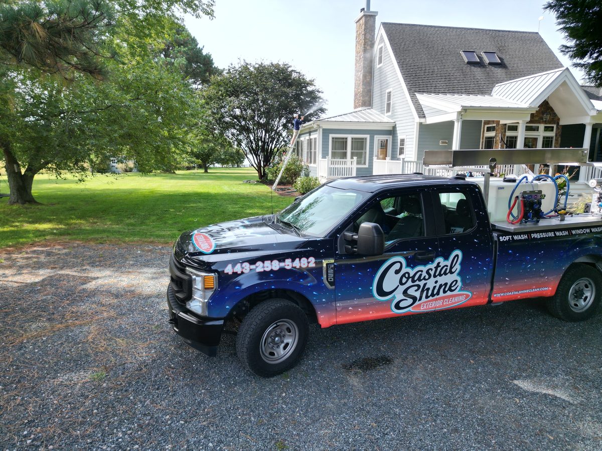 Coastal Shine wrapped work truck in a gravel driveway while a technician soft washes the siding of a gray coastal home