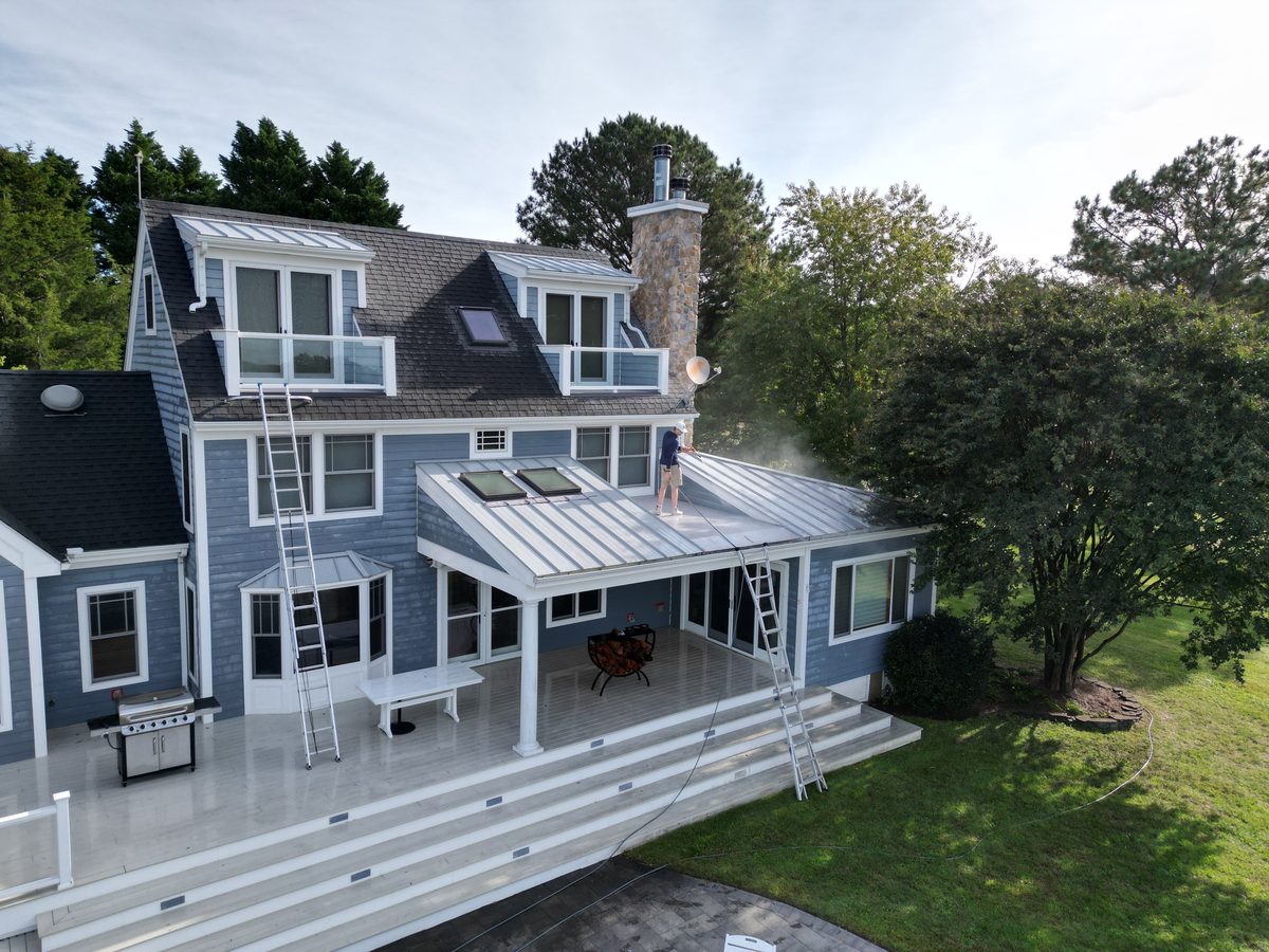Coastal Shine technician on a standing-seam metal porch roof, soft washing the upper siding and dormers of a blue coastal home